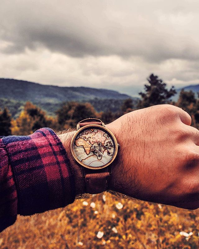 Close-up of a wrist wearing a world map watch, set against a backdrop of mountains and a field, capturing a sense of adventure.