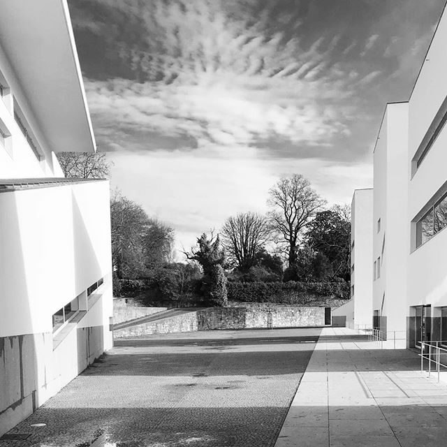 Black and white architectural shot of modern buildings surrounding a courtyard with trees under a cloudy sky.