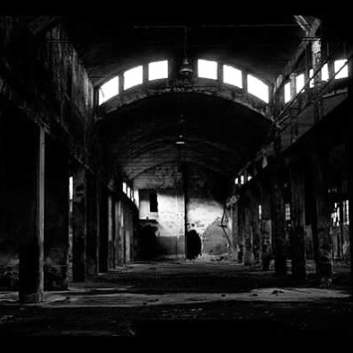 An atmospheric black and white image of an abandoned industrial building interior with pillars and arched windows.