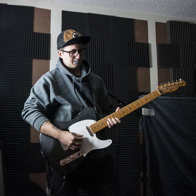 A man wearing glasses and a cap plays an electric guitar in a music studio.