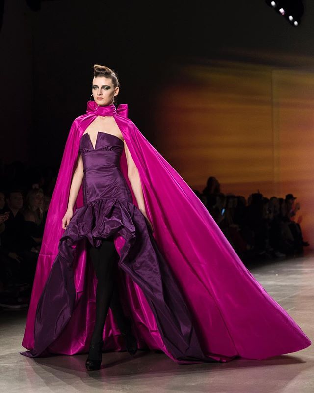 A model walks the runway in a purple gown and magenta cape at a fashion show.