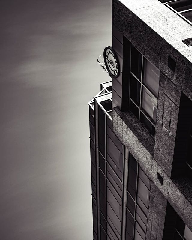 A low-angle, monochrome shot of a modern clock tower and building against a bright sky, emphasizing architectural design.