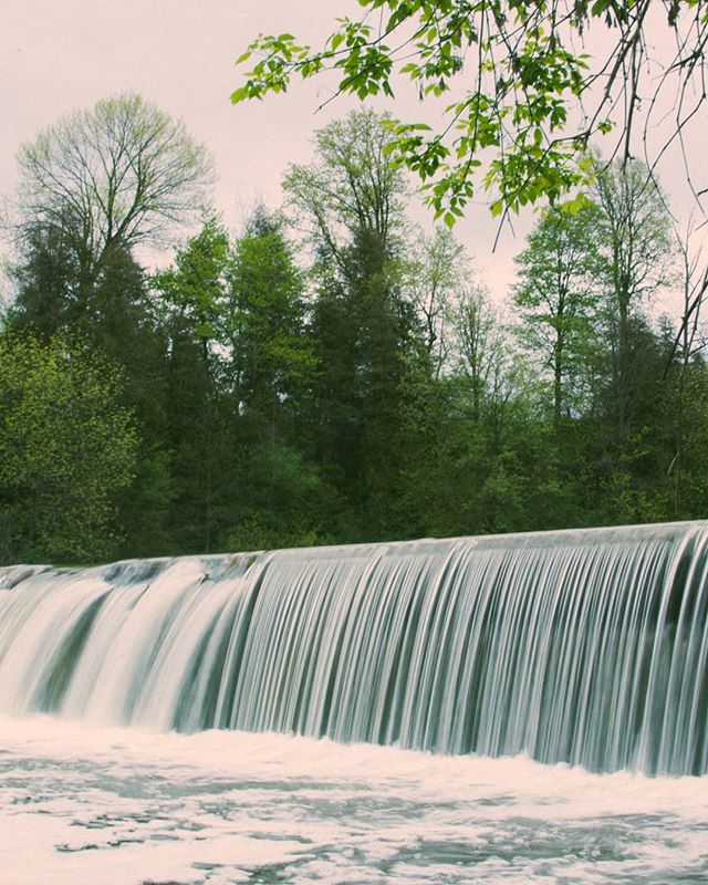 A scenic waterfall flows over a dam, surrounded by lush trees in a calming, nature-filled landscape.