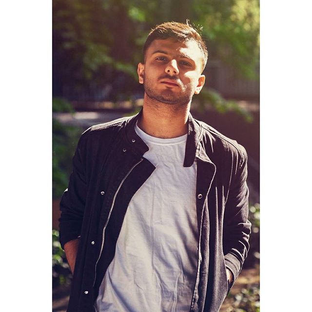A confident young man poses in casual attire outdoors in a modern urban setting.