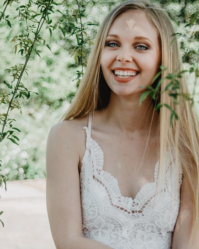 A smiling young woman with blonde hair poses outdoors in a white lace top, surrounded by greenery.