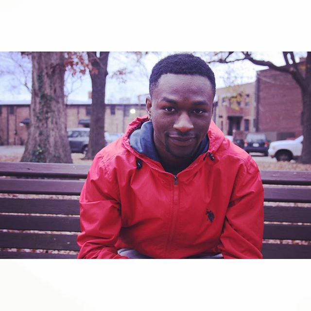 A man in a red jacket sits on a bench in an urban park setting, looking relaxed and friendly.