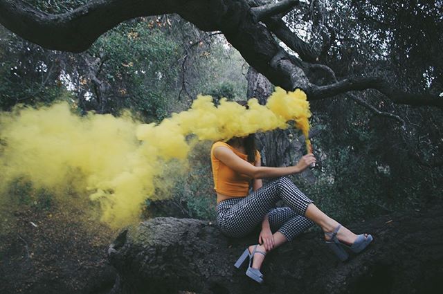 A woman sits on a tree trunk holding a yellow smoke flare in a dark forest setting.