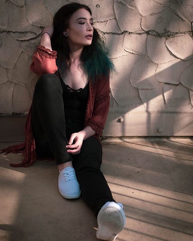 A young woman with dyed hair sits thoughtfully against a textured wall in soft light.