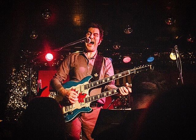 A musician sings and plays a blue double-neck guitar on a brightly lit stage.