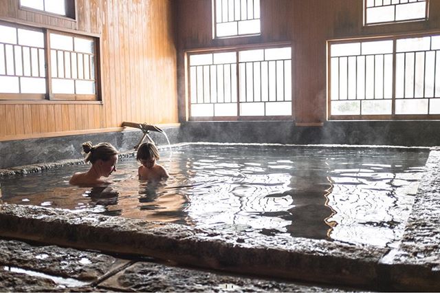 A woman and a child relax in a Japanese onsen with traditional wooden architecture and natural light.