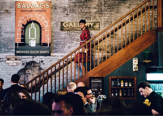 A woman walks down stairs at a busy bar with a vintage atmosphere and many people around.