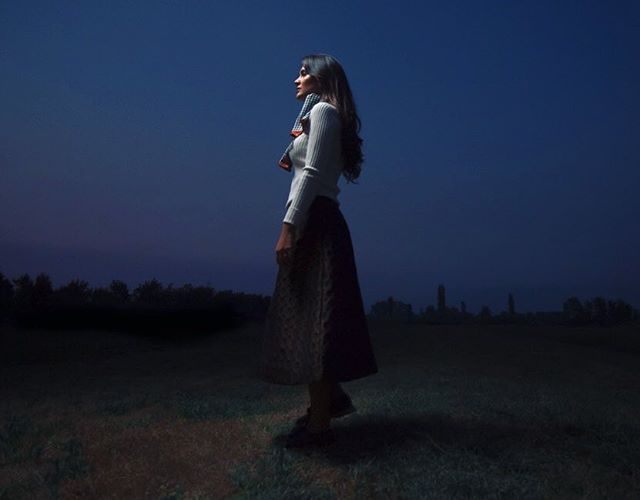 A woman in a dress and scarf stands in a field at night, looking out at the landscape in a dreamy, contemplative scene.