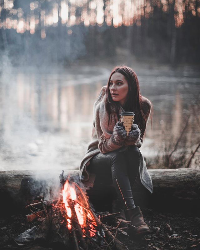 A woman enjoys coffee by a cozy campfire near a serene lake in a peaceful forest setting.