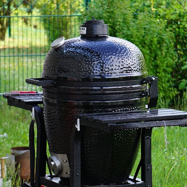 A black kamado grill stands ready in a lush garden setting, promising delicious outdoor meals.