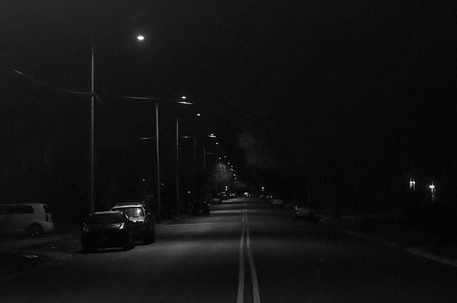 A black and white image depicts a street at night, illuminated by streetlights and lined with parked cars.