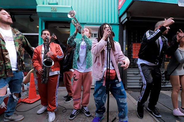 A group of musicians performs on the street, playing instruments with energy and style, surrounded by a small crowd.