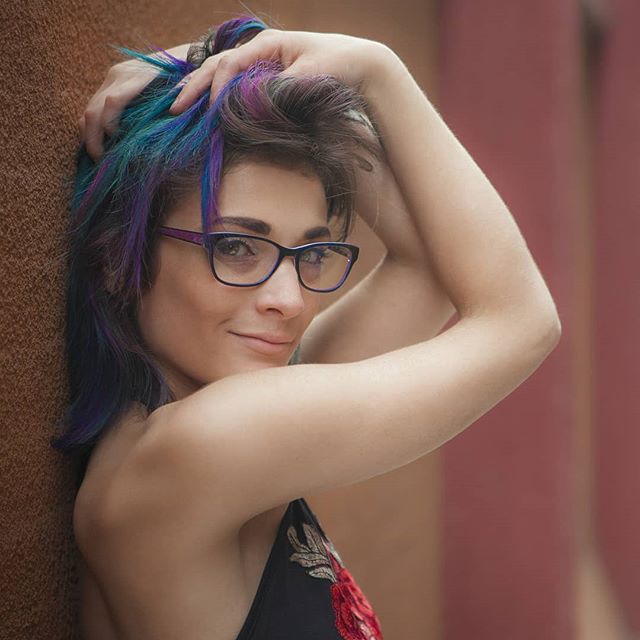 A woman with colorful hair and glasses poses casually, conveying confidence and self-expression.