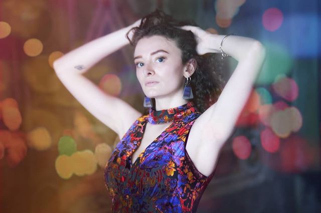 A stylish woman poses with hands behind her head against a bokeh background in a vibrant dress.