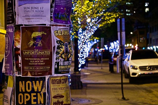 A street pole covered in event posters at night, with decorative lights and a car in the background.