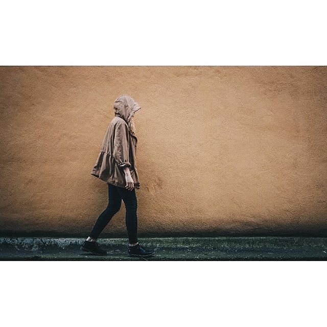 A woman in a hoodie walks along a street against a textured wall.