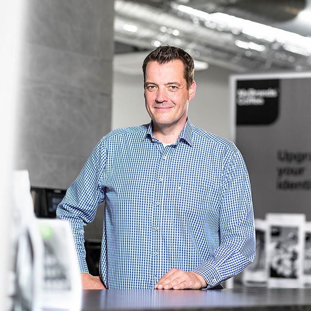 A confident man in a blue shirt stands in an office setting.