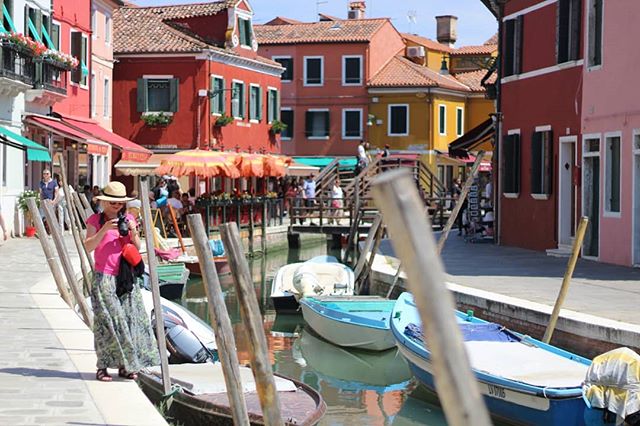 A woman in Burano, Italy stands by the canal with colorful buildings and boats, capturing the scenic landscape.
