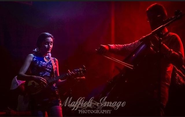 Two musicians are performing passionately on stage, illuminated by vibrant, warm colored stage lights.