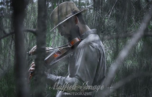 A bearded man in a hat plays the violin in a misty forest, creating a moody and contemplative atmosphere.