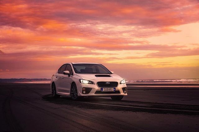 A white Subaru car drives on a dark sand beach during a colorful sunset.