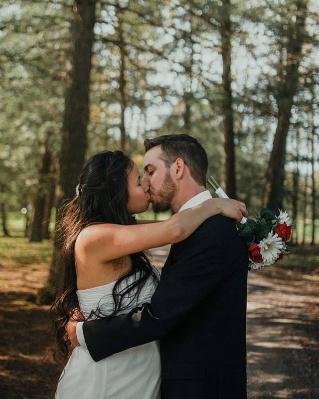 A bride and groom share a tender kiss in a sun-dappled forest, celebrating their wedding day.