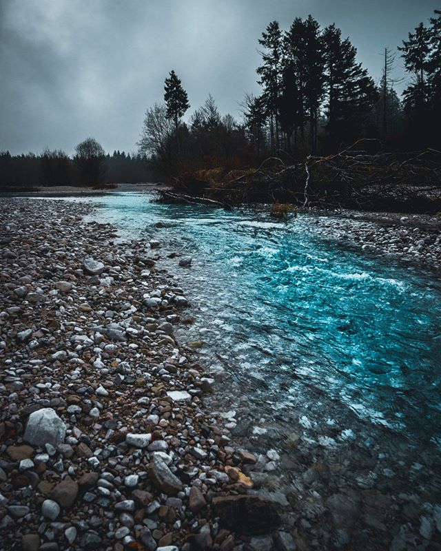 A tranquil river flows through a rocky landscape, surrounded by trees under an overcast sky.