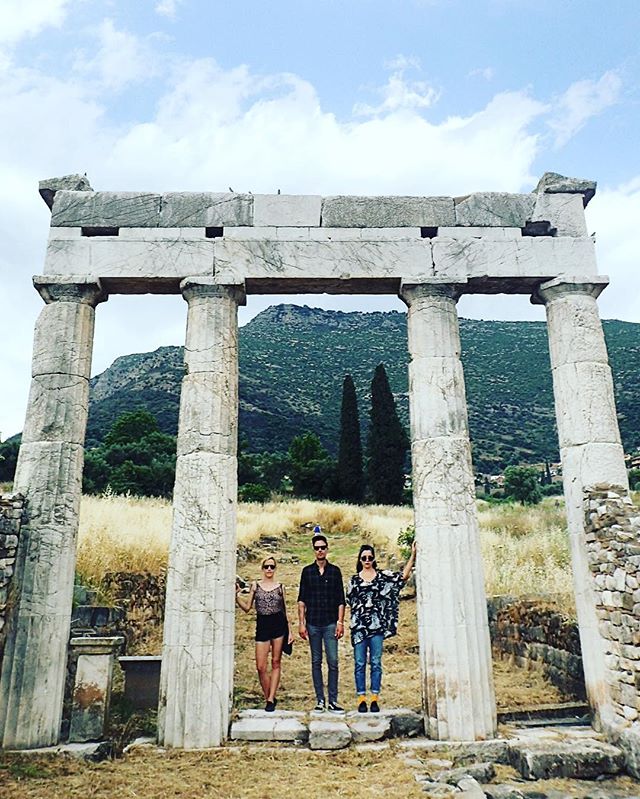 Three people stand under ancient stone columns in Greece, enjoying the historical site and sunny weather.