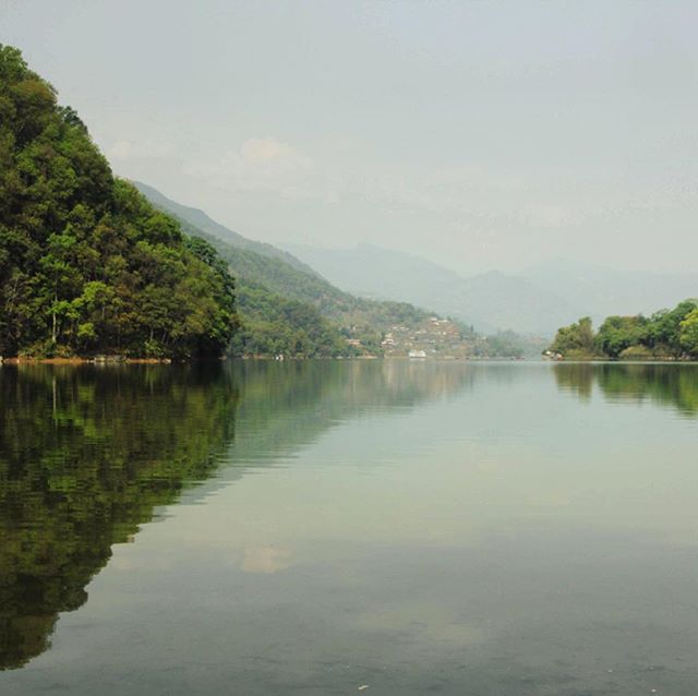 A tranquil lake reflects the surrounding mountains and lush greenery under a serene, overcast sky.