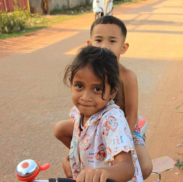 Two cheerful kids are riding a bicycle on a sunny street, looking directly at the camera.