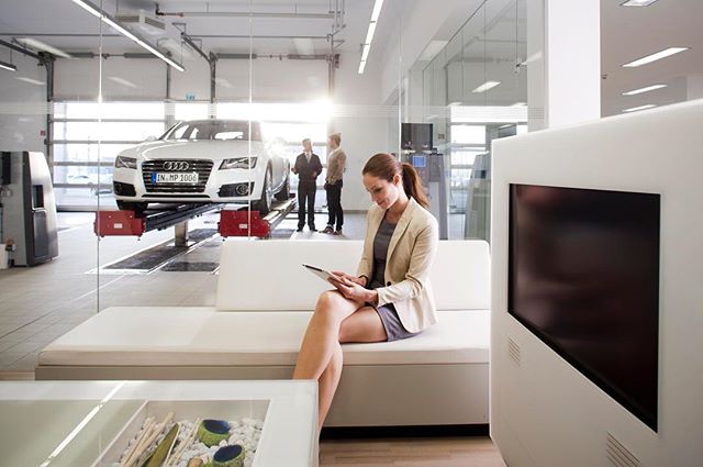 A woman using a tablet sits in a car service waiting room. Men stand near an Audi in the background.
