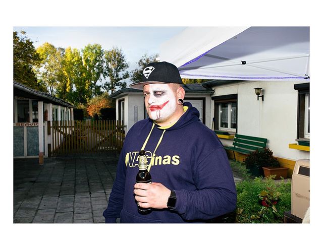 A man in Joker makeup and a Superman hat holds a beer bottle at an outdoor event.