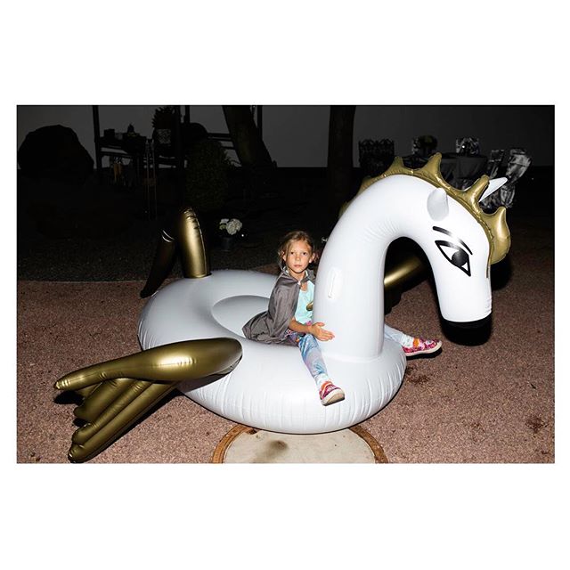 A girl sits on a white and gold inflatable unicorn toy outdoors, enjoying a moment of fun.