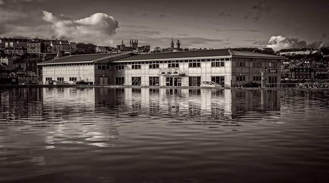 A monochromatic image showcases a building near a body of water with reflections and a cityscape in the background.