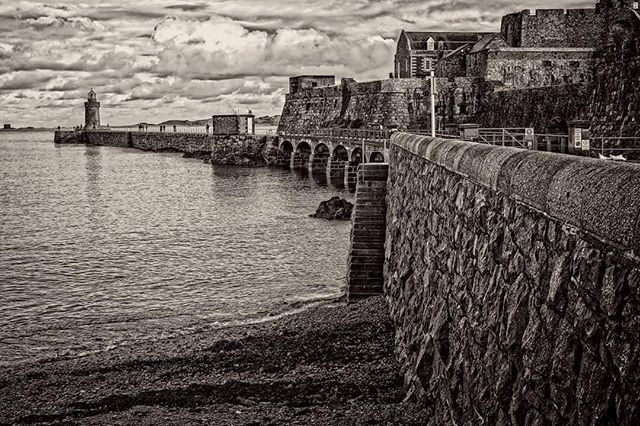 A monochrome image of a historic stone fortress and lighthouse along a coastline under a cloudy sky.
