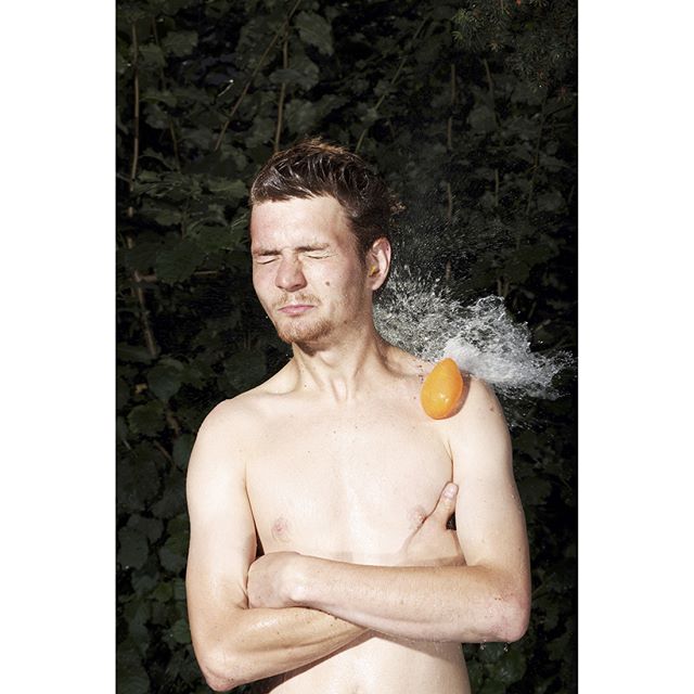 A shirtless man is hit by a water balloon outdoors, a playful, summer scene.