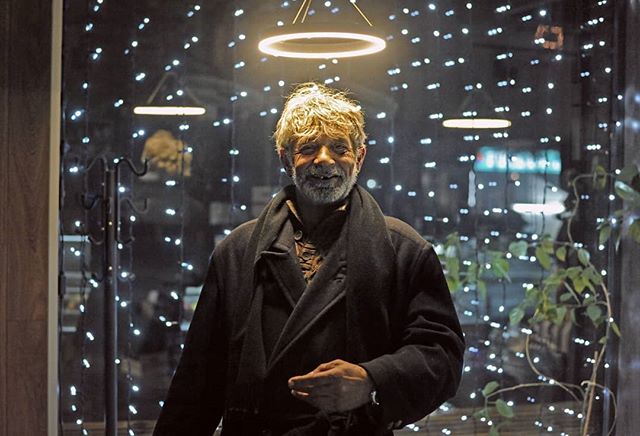 A smiling man in a coat stands indoors with lights creating a warm and inviting atmosphere.