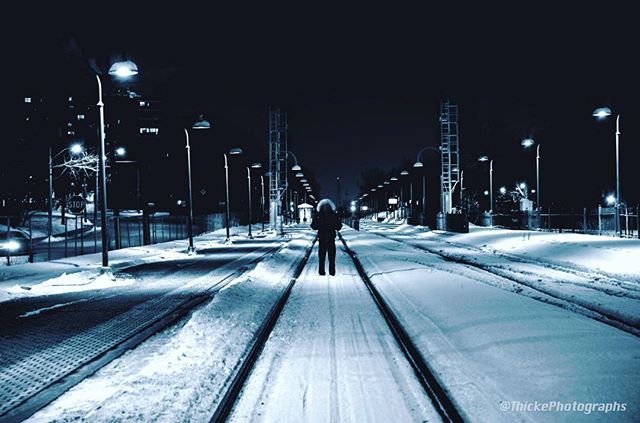 A lone figure walks along snowy train tracks at night, surrounded by streetlights.