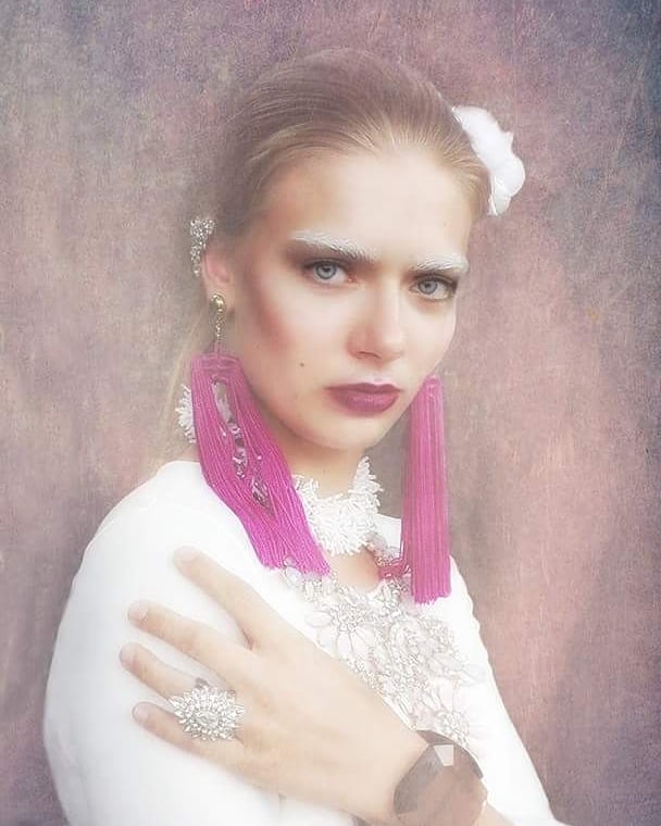 A woman with white eyebrows poses in an editorial style shoot with pink tassel earrings, and a lacy white dress.