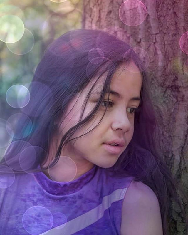 A young girl with dark hair and a purple shirt gazes softly against a tree in a dreamy, bokeh-filled outdoor setting.