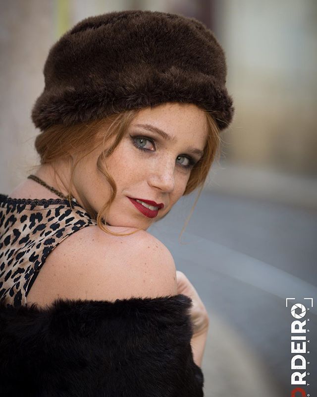 A redhead woman in a vintage style hat and leopard print top looks over her shoulder with a mysterious expression.