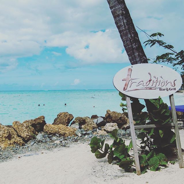 A scenic view of a beach with a Traditions Bar & Grill sign, rocks, and the ocean under a blue sky.