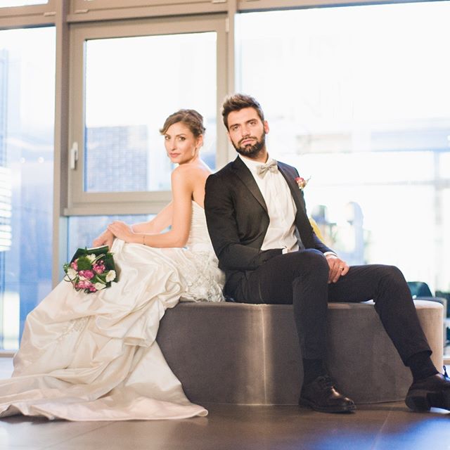 A bride and groom are sitting back to back, posing for their wedding photos in front of a window.