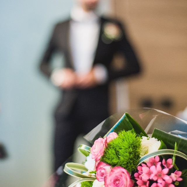 A groom in a tuxedo stands behind a beautiful bouquet of pink, white, and green flowers.