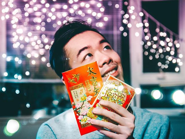 A smiling man holds red envelopes against a backdrop of soft, festive lights.