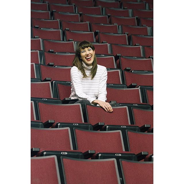 A happy woman sits in a red theater seat, smiling at the camera.